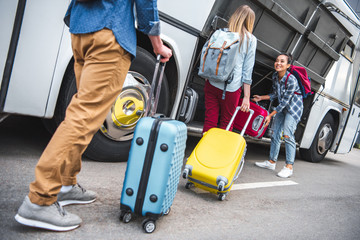 selective focus of smiling woman putting wheeled bag into travel bus while her friends walking near at urban street