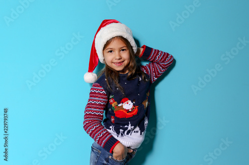 Cute little girl in Christmas sweater and hat on color background