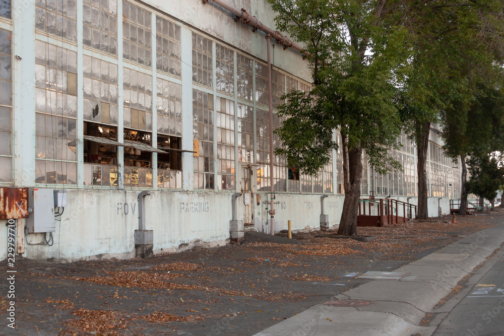 The white painted side of a WW II industrial building. It has tiers of windows that open horizontally. Trees are along the front. Parking spaces are in front of the building.