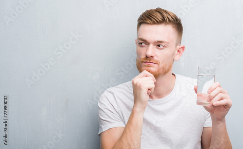 Young redhead man over grey grunge wall drinking water serious face thinking about question, very confused idea