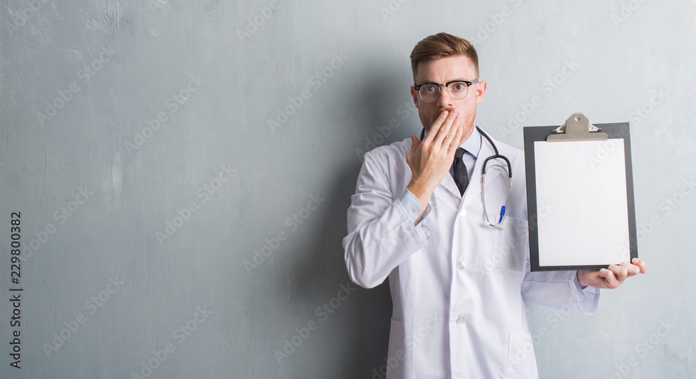 Young redhead doctor man over grey grunge wall holding clipboard cover mouth with hand shocked with shame for mistake, expression of fear, scared in silence, secret concept Young redhead doctor man over grey grunge wall holding clipboard cover mouth with hand shocked with shame for mistake, expression of fear, scared in silence, secret concept
