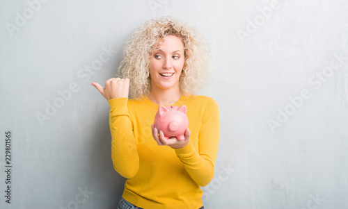 Young blonde woman over grunge grey background holding piggy bank pointing and showing with thumb up to the side with happy face smiling