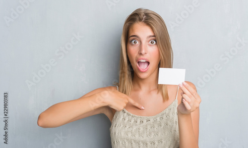 Beautiful young woman over grunge grey wall holding blank visit card with surprise face pointing finger to himself