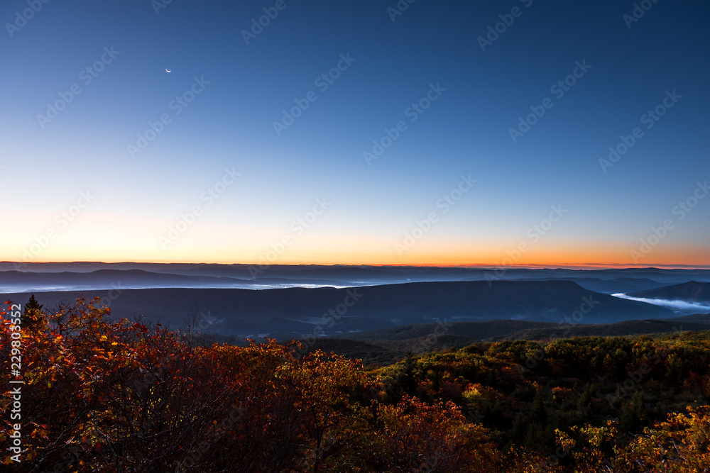 Bear rocks overlook during sunrise, dawn, moon in autumn with rocky landscape in Dolly Sods, West Virginia with orange foliage trees, blue, yellow sky, layered mountains, hills