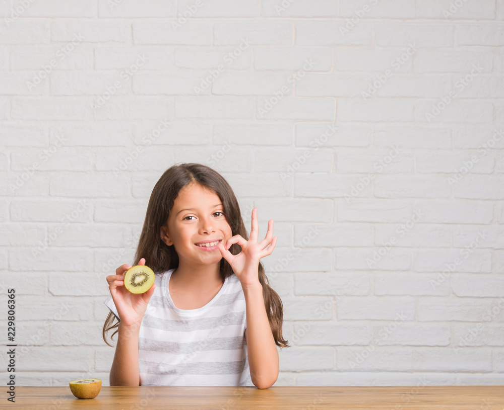 Young hispanic kid sitting on the table eating kiwi doing ok sign with fingers, excellent symbol Young hispanic kid sitting on the table eating kiwi doing ok sign with fingers, excellent symbol