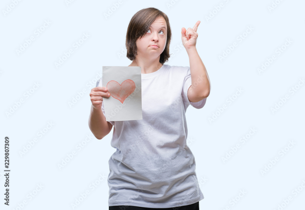 Young adult woman with down syndrome holding red heart card over isolated background very happy pointing with hand and finger to the side Young adult woman with down syndrome holding red heart card over isolated background very happy pointing with hand and finger to the side