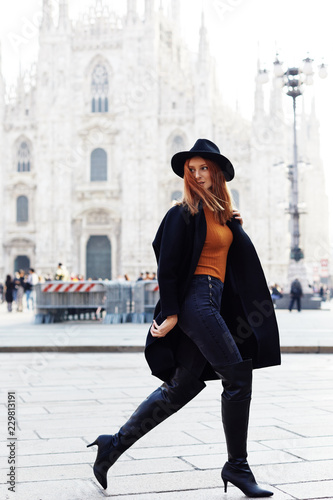 Fashion model walking by the Duomo of Milan wearing a hat and a dark coat in a October morning looking back and movement on the hair
