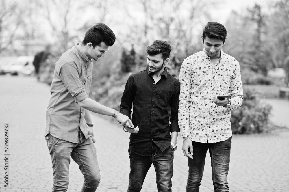 Three indian guys students friends walking on street and looking at mobile phone. Black and white photo. Three indian guys students friends walking on street and looking at mobile phone. Black and white photo.