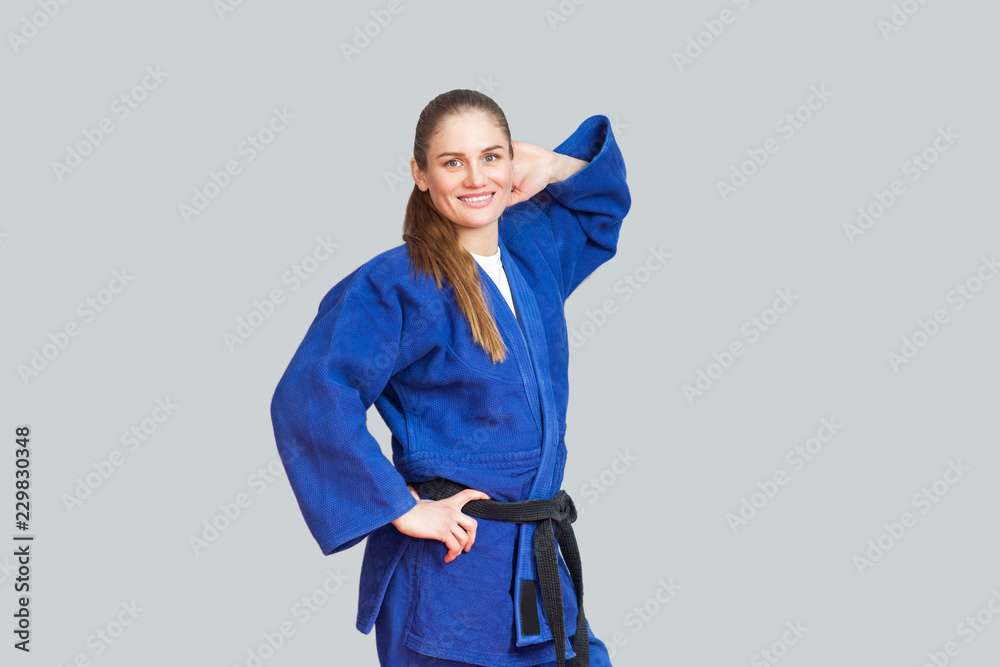 Beautiful happy athletic karate woman in blue kimono with black belt posing  and holding hand behind her head are looking at camera. Japanese martial  arts concept. Indoor, studio shot, gray background Stock-Foto