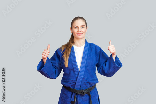 Positive athletic karate woman in blue kimono with black belt standing  showing thumbs up and looking at camera with toothy smile. Japanese martial arts concept. Indoor  studio shot  gray background