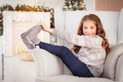 Merry Christmas and Happy New Year. A beautiful, little girl funny sitting on a chair in the New Year's environment. A child in a knitted sweater and knitted boots. Snow, a fairy tale, a miracle.