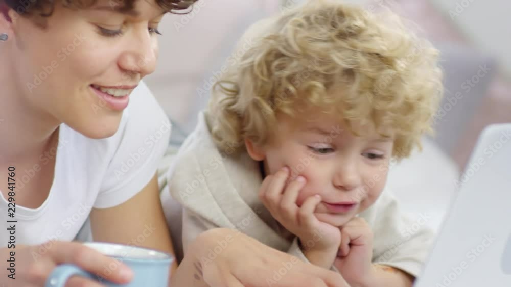 Medium shot of happy curly mother and adorable little boy lying on bed on weekend and chatting while doing something on laptop Medium shot of happy curly mother and adorable little boy lying on bed on weekend and chatting while doing something on laptop