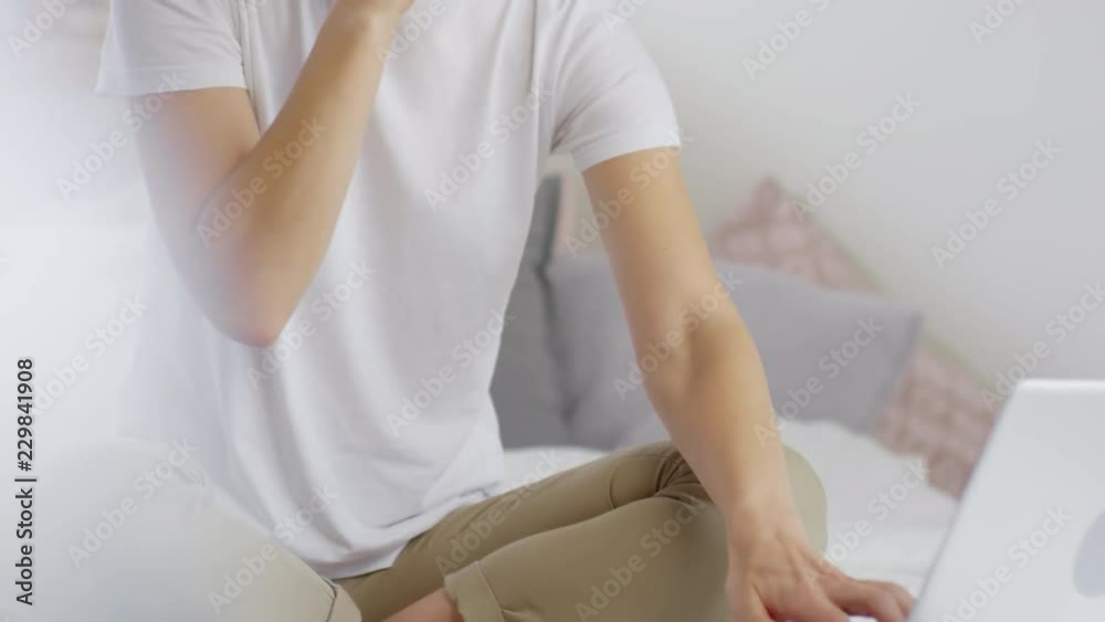 Handheld tilt up shot of cheerful woman with short curly hair sitting on bed and making phone call while working on laptop in morning Handheld tilt up shot of cheerful woman with short curly hair sitting on bed and making phone call while working on laptop in morning
