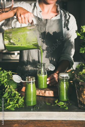 Making green detox take-away smoothie. Young female in linen apron pouring green smoothie drink from blender to bottle surrounded with vegetables and greens. Healthy, weight loss food concept