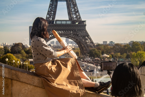 Woman holding a french baguette posing at the Trocadero stairs with the Eiffel Tower on the background on a sunny day with blue sky and clouds in Paris, France while woman take a photo with smartphone