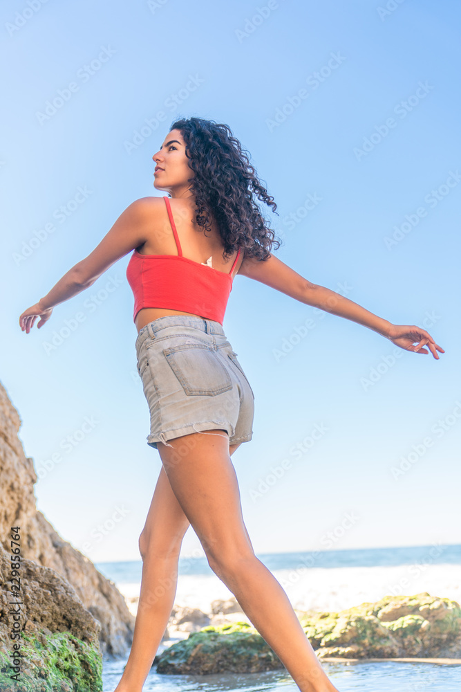 Portrait of latino woman with curly hair, red top and denim shorts running at beach Portrait of latino woman with curly hair, red top and denim shorts running at beach