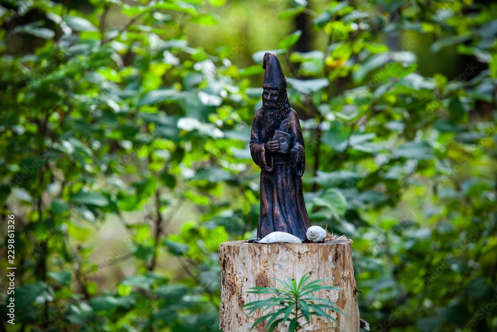 Statue representing a magicien holding a sacred magic book, displayed on a tree stomp with eggshells and a blurry background of green bushes