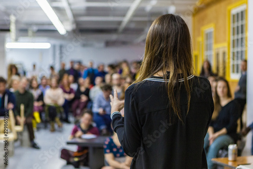 Young woman is giving a conference in front of 200 people in an industrial environment - back view - Blurred audience mainly composed of young adults
