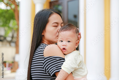 Portrait of Asian mother carrying and kissing her infant baby boy outdoor.
