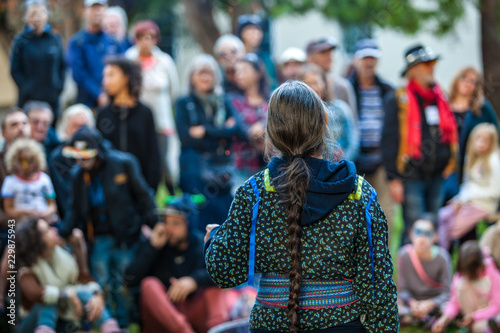 Woman with platted hair and glasses is giving a public speech or conference at the park in front of a blurry crowd of 50 people photo