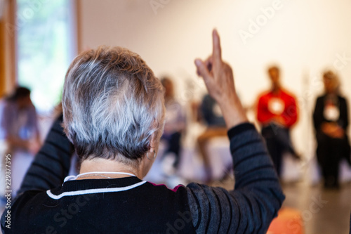 Woman with short black and white hairs and raising both of her arms is talking in front of a full committee - Pictured from the back in an alternative health center photo