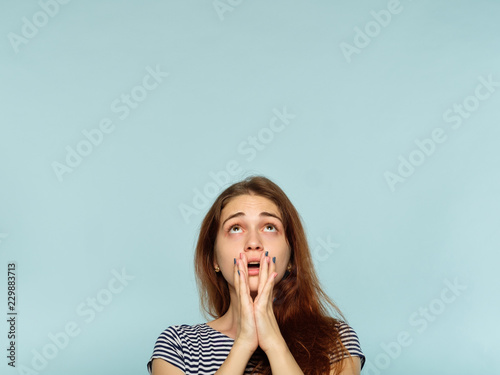 emotion face. overwhelmed perplexed shocked surprised astounded woman looking up and above. young beautiful brown haired girl portrait on blue background.