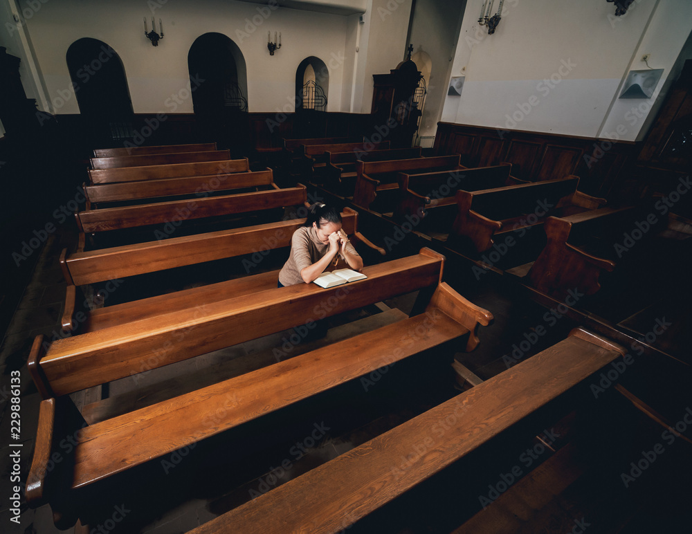 Christian woman praying in church. Hands crossed and Holy Bible on wooden desk.