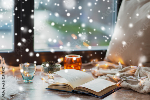 hygge, christmas and cozy home concept - book, garland lights and candles on window sill over snow