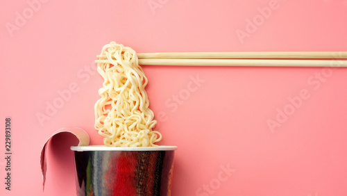 Instant noodles cup and chopsticks on a pink background.