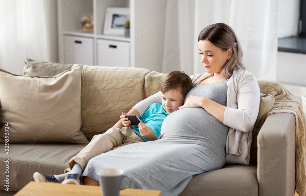 family, technology and pregnancy concept - happy pregnant mother and little son with smartphone at home