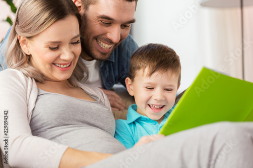 pregnancy, people and family concept - happy pregnant mother, father and little son reading book on sofa at home