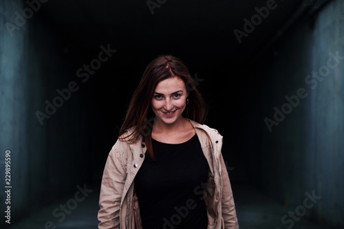 Lifestyle image of a young pretty chestnut haired caucasian girl walking out of dark concrete corridor smiling . Cute casual outfit. photo