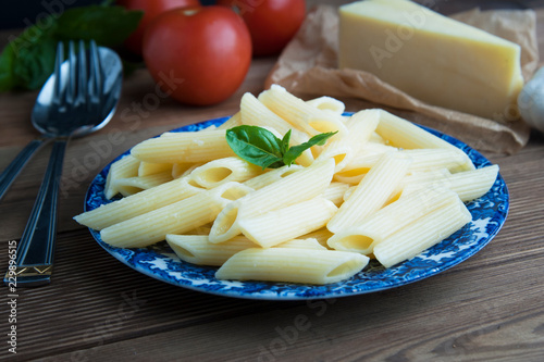 Cooked penne pasta with butter,cheese and fresh vegetable ingredients on background: tomato, garlic, onion, fork and spoon, basil. Vintage beautiful blue plate on wooden table. Isolated.