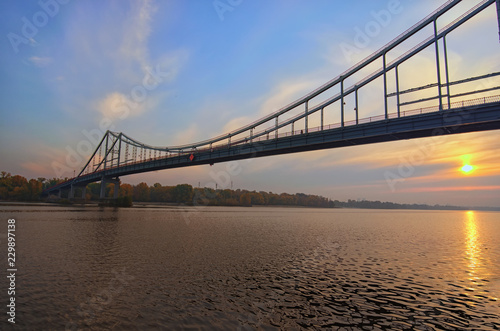 Wide angle landscape panorama of Dnipro River and Pedestrian Bridge to the Trukhanov Island. Colorful vibrant sky, sun reflected in the water. Foggy autumn morning during sunrise. Kyiv, Ukraine