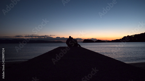 A couple sit back to back on a sea pier at sundown to create a beautiful romantic photo. The 2 people are silhouetted against a sunset sky and a calm ocean. 