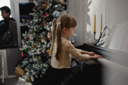 Girl is playing the piano at the Christmas tree with shadows around and a man silhouette at the window