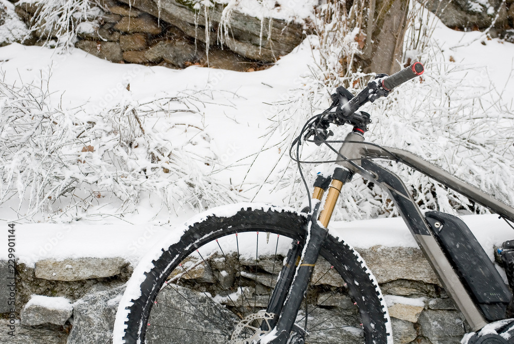detail of electric bicycle, e-bike, ebike, resting on a wall of rocks, mountain forest during snow storm, blizzard, sports, end of winter, handlebars, wheel, display, cold, alps, Piedmont, Italy detail of electric bicycle, e-bike, ebike, resting on a wall of rocks, mountain forest during snow storm, blizzard, sports, end of winter, handlebars, wheel, display, cold, alps, Piedmont, Italy