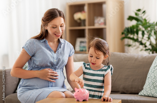 pregnancy, finances and family concept - happy pregnant mother and little daughter putting euro coin into piggy bank at home