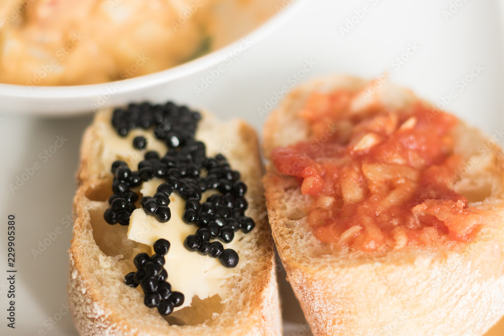 Close-up: two open sandwiches on a plate. One piece of bread is with butter and black caviar. Another piece of bread with tomato, onion and garlic.