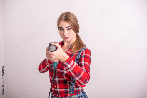 Young sexy woman stands near the white wall and holds a drill. Girl in shorts and a plaid shirt takes aim with a puncher. A woman makes repairs with a tool.