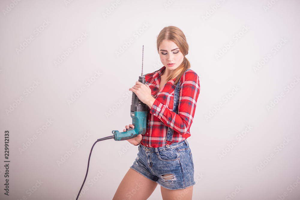 Young sexy woman stands near the white wall and holds a drill. The girl in shorts and a plaid shirt works as a puncher. A woman makes repairs with a tool. Young sexy woman stands near the white wall and holds a drill. The girl in shorts and a plaid shirt works as a puncher. A woman makes repairs with a tool.