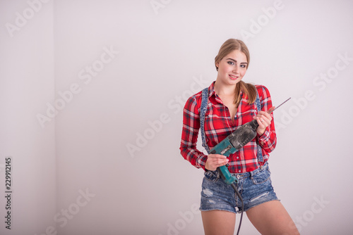 Young sexy woman stands near the white wall and holds a drill. The girl in shorts and a plaid shirt works as a puncher. A woman makes repairs with a tool.