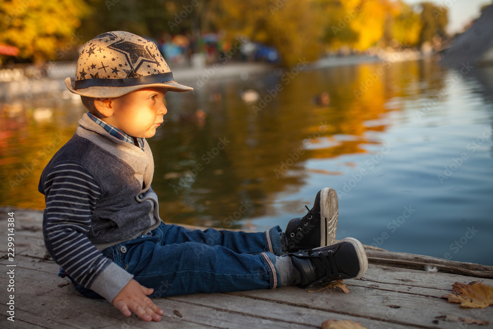 A little boy is sitting next to the lake during a walk in the park in autumn. A little boy is sitting next to the lake during a walk in the park in autumn.