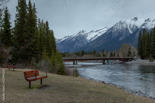 Public bench on the bank of the Bow River with the view of the bridge to downtown Canmore and the Canadian Rocky Mountains in Alberta