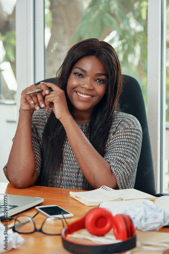 Obraz premium Happy adult black woman sitting at working desk in office smiling at camera adorably