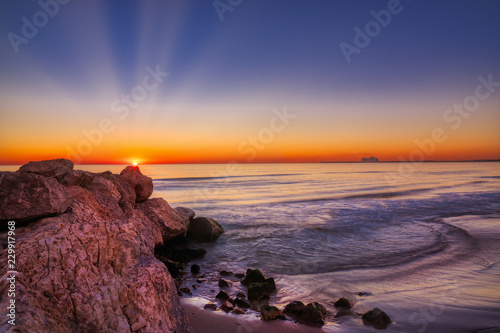 A deep and colorful sunrise over rocks and a sandy sea shore with blues, reds, oranges and yellows, and a cruise ship in the far distance docked at a port
