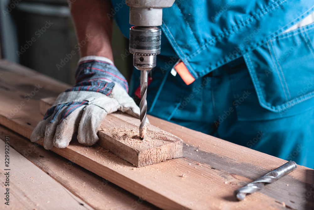A man joiner in overalls and gloves drills a wooden Board close-up.