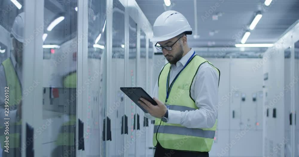 Adult man in hardhat walking in control center of solar electric plant using tablet and checking electrical engineering behind glass door Adult man in hardhat walking in control center of solar electric plant using tablet and checking electrical engineering behind glass door