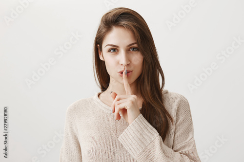 Keep it information in secret. Indoor shot of attractive sensual brunette in beige sweater smiling curiously and showing shh or shush gesture with index finger over mouth, wanting to hide something