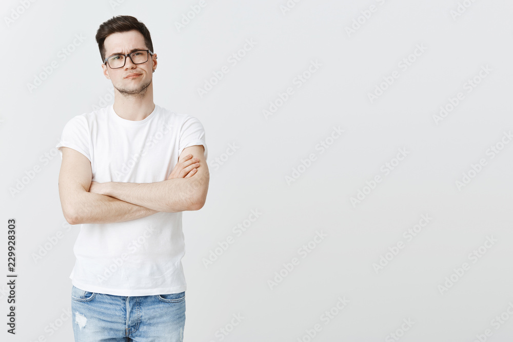 Guy standing with displeased look disliking, making negative opinion about show. Disappointed young attractive man with bristle in glasses crossing hands on chest and smirking from dissatisfaction